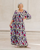 A woman wearing a long-sleeved, floor-length floral maxi dress with a black base and pink, purple, and white flowers, standing on stone steps.