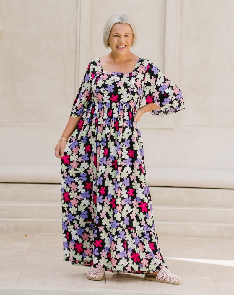 A woman wearing a long-sleeved, floor-length floral maxi dress with a black base and pink, purple, and white flowers, standing on stone steps.