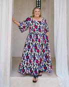 A woman modeling a long-sleeve, floor-length floral maxi dress with a black background and pink, purple, and white flowers, standing in a doorway with white drapes.