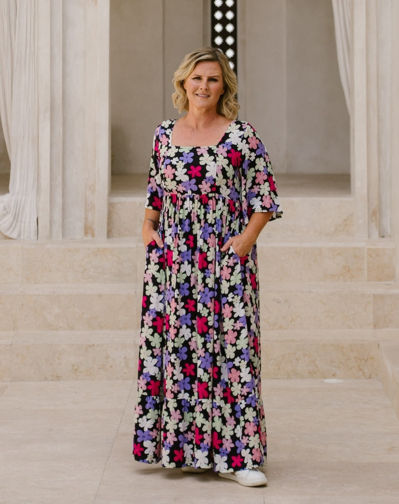 A woman wearing a long-sleeved, knee-length maxi dress with a colorful floral print stands on stone steps in front of a classical building with columns.