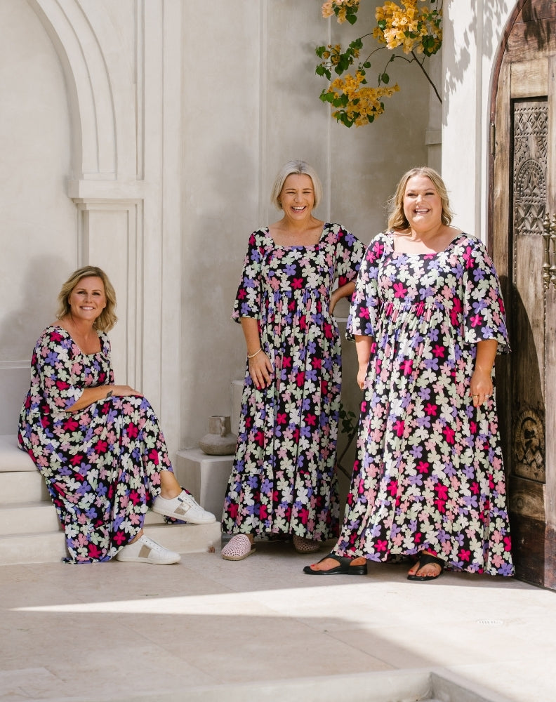 Three women wearing matching floral maxi dresses in a sunlit outdoor courtyard with white arches and a wooden door. The dresses have a dense floral print in pink, purple, blue, and white on a light base.