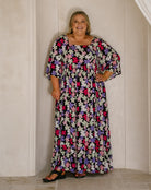 A woman modeling a long-sleeved, floor-length maxi dress with a dense multicolor floral print on a black background, standing against a cream-colored paneled wall.