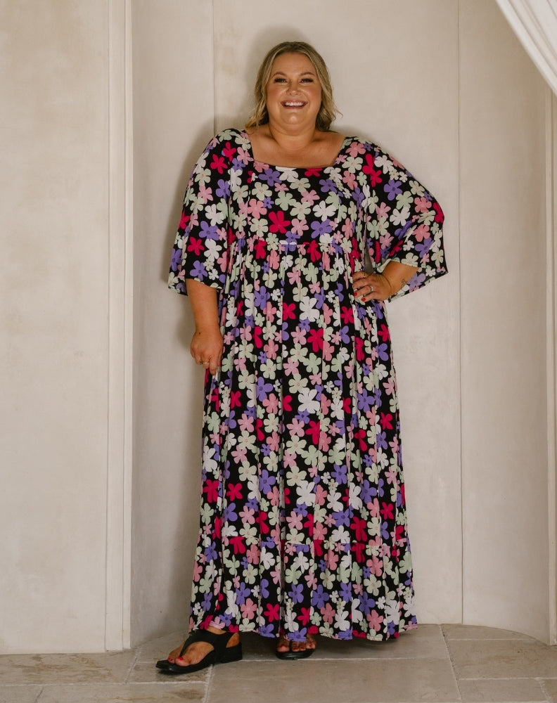 A woman modeling a long-sleeved, floor-length maxi dress with a dense multicolor floral print on a black background, standing against a cream-colored paneled wall.