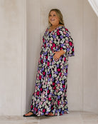 A woman wearing a long-sleeve, floor-length floral maxi dress with a black base and pink, purple, and white flowers, standing in a tiled room with beige walls.