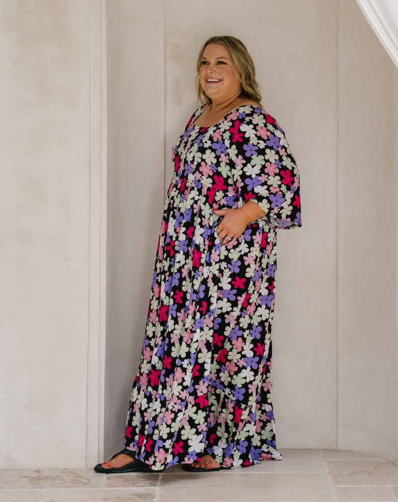 A woman wearing a long-sleeve, floor-length floral maxi dress with a black base and pink, purple, and white flowers, standing in a tiled room with beige walls.