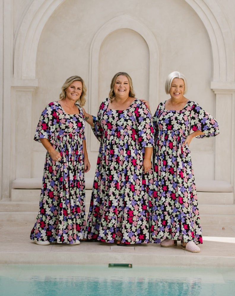 Three women wearing matching floral maxi dresses stand together in front of a white stone archway, with a pool of water visible in the foreground.