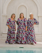 Three women wearing matching floral maxi dresses stand together in front of a white stone archway, with a pool of water visible in the foreground.