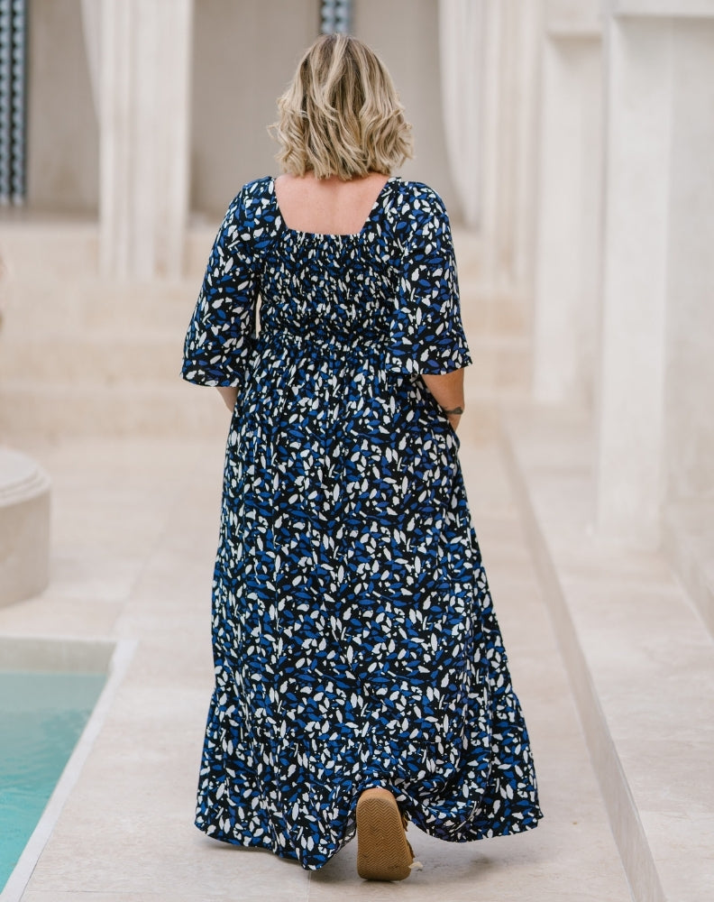 A woman viewed from behind wearing a navy blue maxi dress with a white abstract floral pattern, walking on a light stone walkway next to a pool.