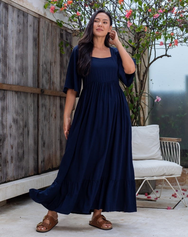 Woman wearing a navy blue dress standing outdoors with a wooden fence and white chair in the background.