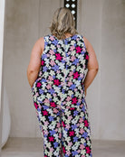 A woman viewed from behind wearing a sleeveless jumpsuit with a dense multicolor floral print on a black background. The print features white, pink, and purple flowers.