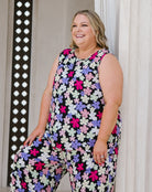 A woman wearing a sleeveless jumpsuit with a colorful floral pattern, standing in front of a white curtain.