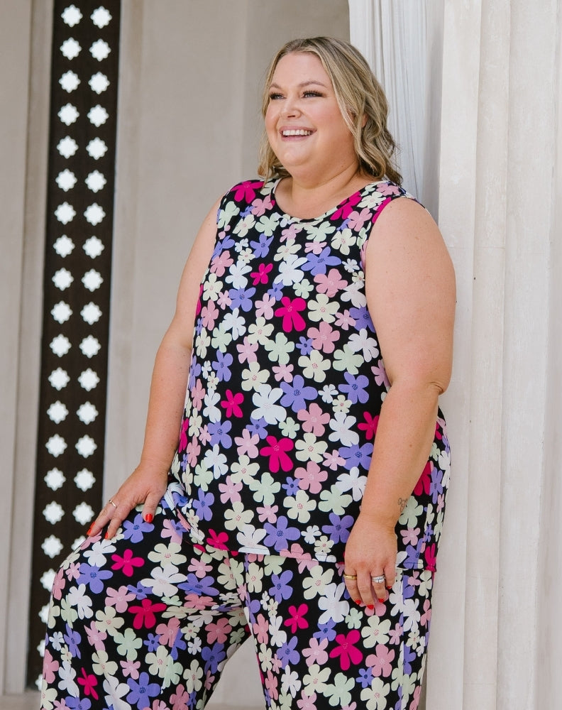 A woman wearing a sleeveless jumpsuit with a colorful floral pattern, standing in front of a white curtain.
