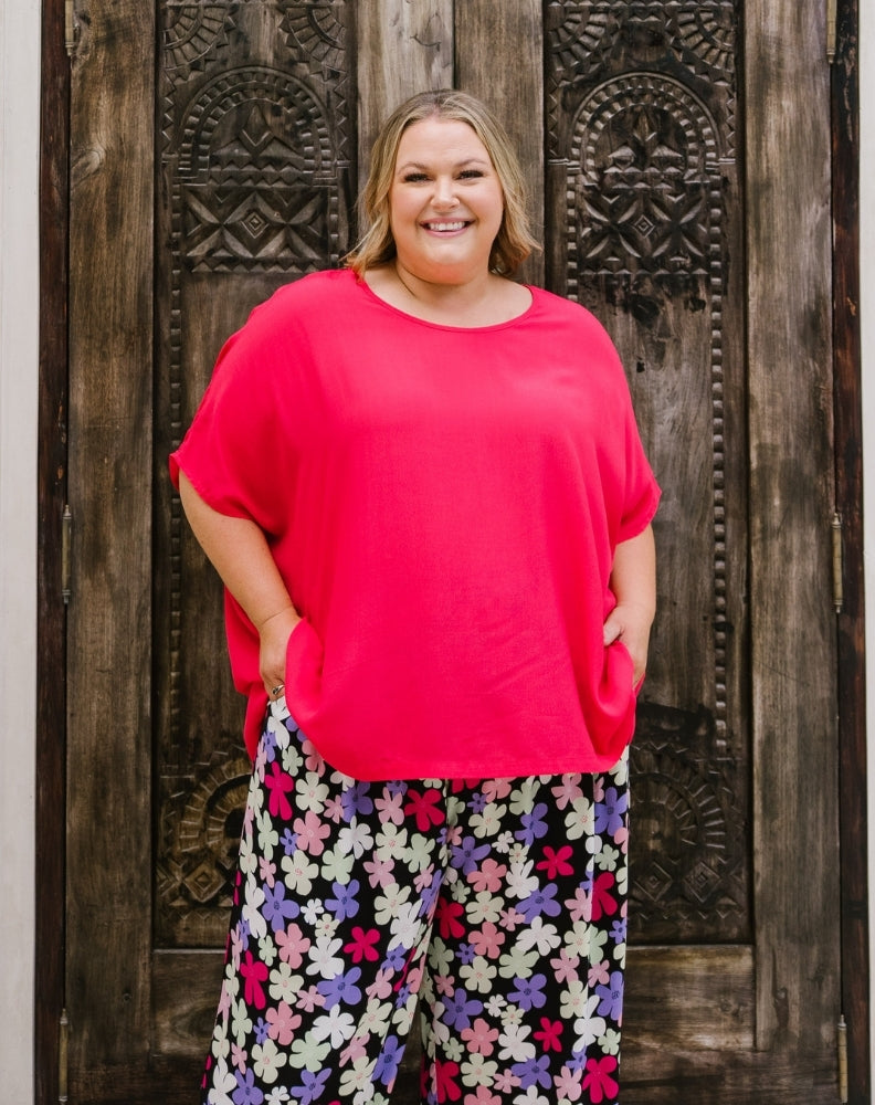A woman stands in front of a dark carved wooden door wearing a bright pink short-sleeve top and black wide-leg pants with a colorful floral print.