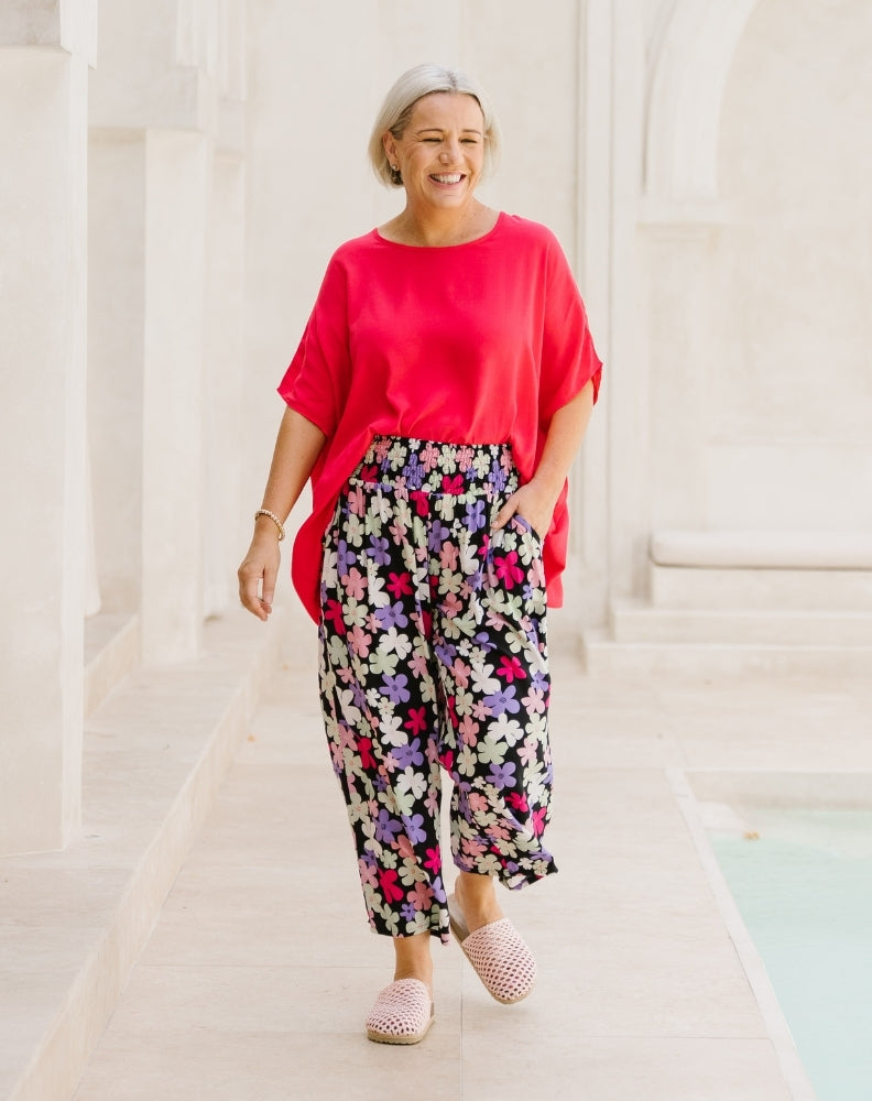A woman walking in a bright, white architectural corridor wearing a bright pinkish-red short-sleeve top, black wide-leg pants with a colorful floral print, and pink slip-on shoes.