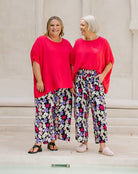 Two women standing side by side wearing matching bright pink short-sleeve tops and wide-leg floral pants with black, white, purple, and pink flowers on a light beige background.