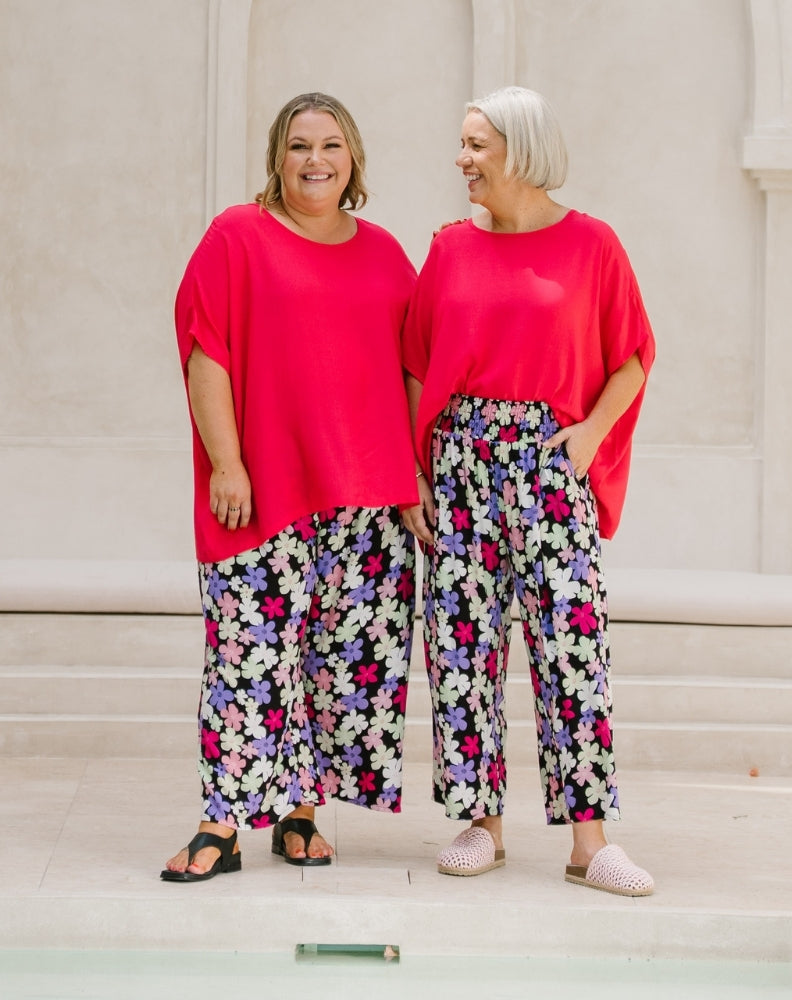 Two women standing side by side wearing matching bright pink short-sleeve tops and wide-leg floral pants with black, white, purple, and pink flowers on a light beige background.