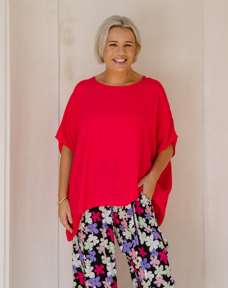 A smiling woman with short blonde hair wearing a bright red oversized short-sleeve top and black pants with a colorful floral print, standing against a light beige wall.