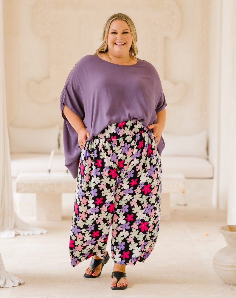 A woman modeling a purple short-sleeve top and high-waisted, wide-leg harem pants with a colorful floral print in pink, purple, white, and black, set against a bright, neutral indoor background.