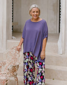 A smiling woman stands on stone steps wearing a loose purple top and colorful floral wide-leg pants, holding a white ornate chair beside her.