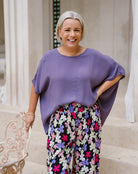 A smiling woman with short blonde hair stands on stone steps wearing a loose, draped lavender-purple top and colorful floral wide-leg pants. She is posed next to a decorative white chair in an outdoor architectural setting.
