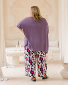 A woman viewed from behind wearing a loose purple top and black wide-leg pants with a colorful floral print, standing in a bright, neutral-toned indoor setting.