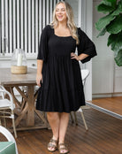Woman wearing a black dress standing on a wooden deck with a table and chairs in the background.