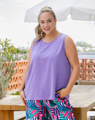 A woman wearing a sleeveless lavender-purple top and colorful geometric print pants, standing outdoors under a white patio umbrella.