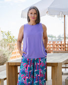 A woman modeling a sleeveless lavender-purple top and colorful geometric print pants on an outdoor patio with a wooden table and umbrella in the background.