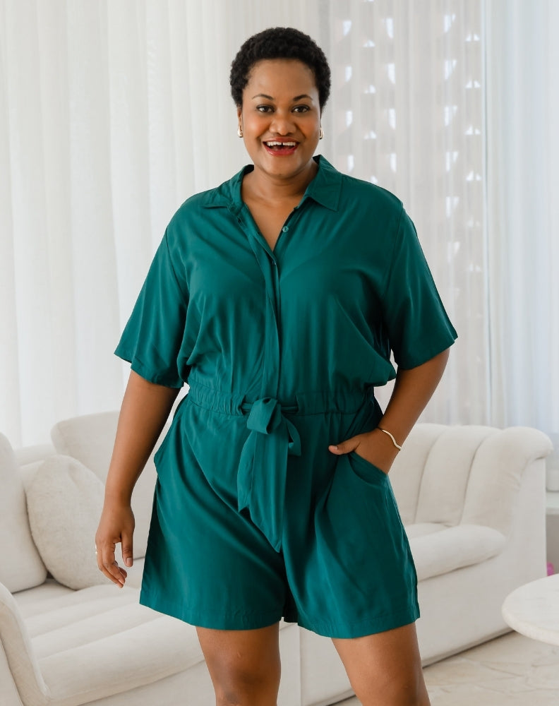 Woman wearing a teal romper standing in a room with white curtains and a white couch.