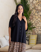 Woman wearing a black shirt and leopard print pants standing in a living room with a stone wall and plants.