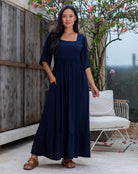 Woman wearing a navy blue dress standing outdoors with a wooden fence and white chair in the background.