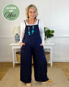 Woman wearing navy blue overalls standing in a room with a white wall and wooden floor.