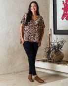 Woman wearing a leopard print top and black pants standing in a room with a decorative plant and artwork.