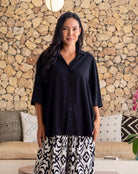 Woman wearing a black shirt and patterned pants standing in front of a stone wall.