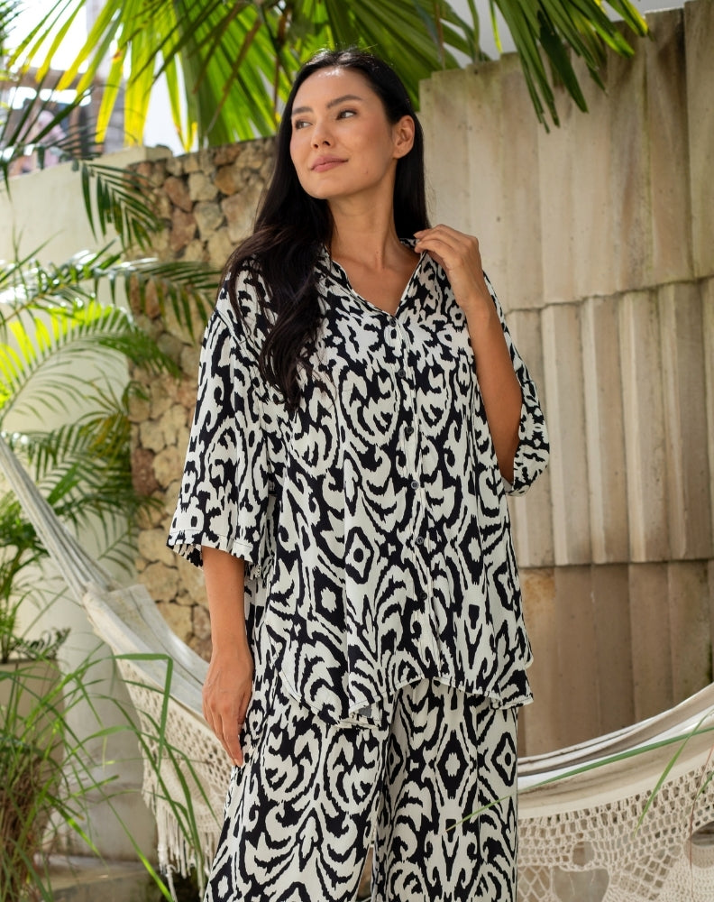 Woman wearing a black and white patterned dress standing outdoors with plants and a wooden wall in the background.