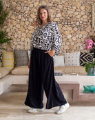 Woman in a patterned top and black pants standing in a living room with a stone wall background.