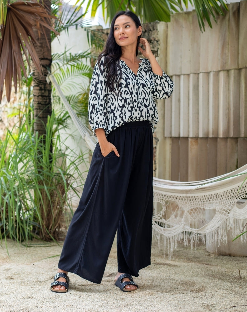 Woman wearing a black and white patterned blouse and dark blue pants standing in a garden setting.