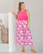 Woman wearing a pink sleeveless top and patterned pants standing against a neutral wall.