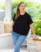 A woman wearing a black short-sleeve t-shirt and light blue jeans stands on a covered patio with green garden foliage in the background.
