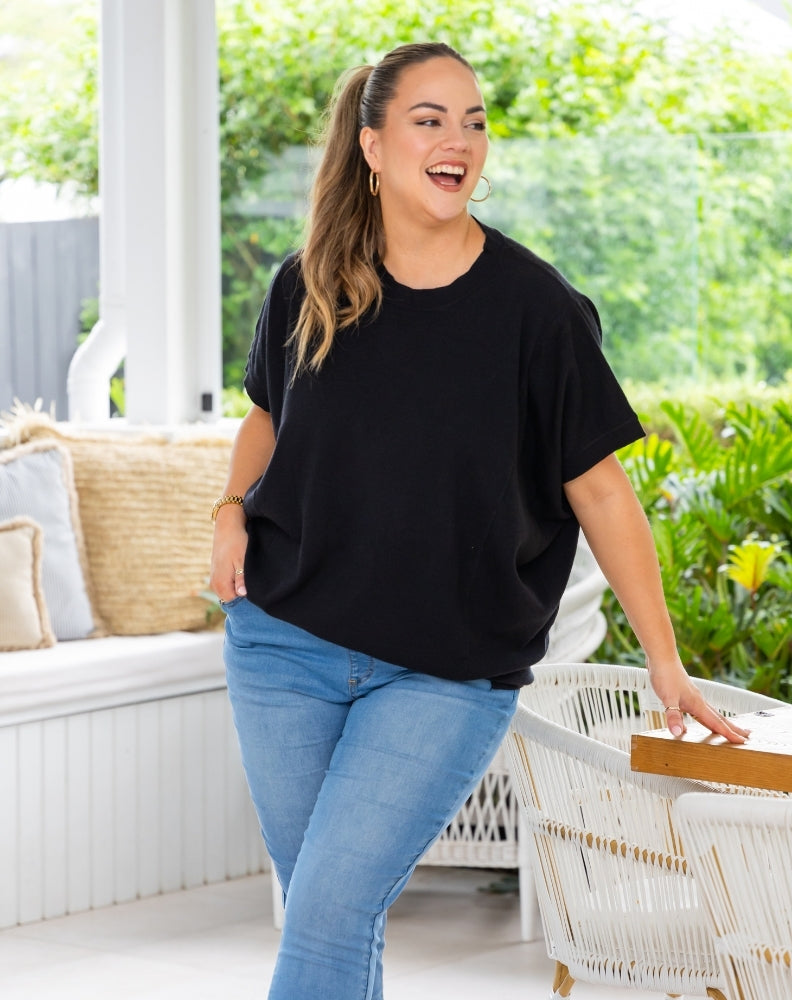 A woman wearing a black short-sleeve t-shirt and light blue jeans stands on a covered patio with green garden foliage in the background.