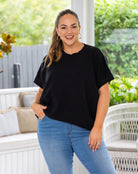 A woman modeling a black short-sleeve crewneck top with scalloped edges, paired with light blue jeans, standing on a white patio with green garden foliage in the background.