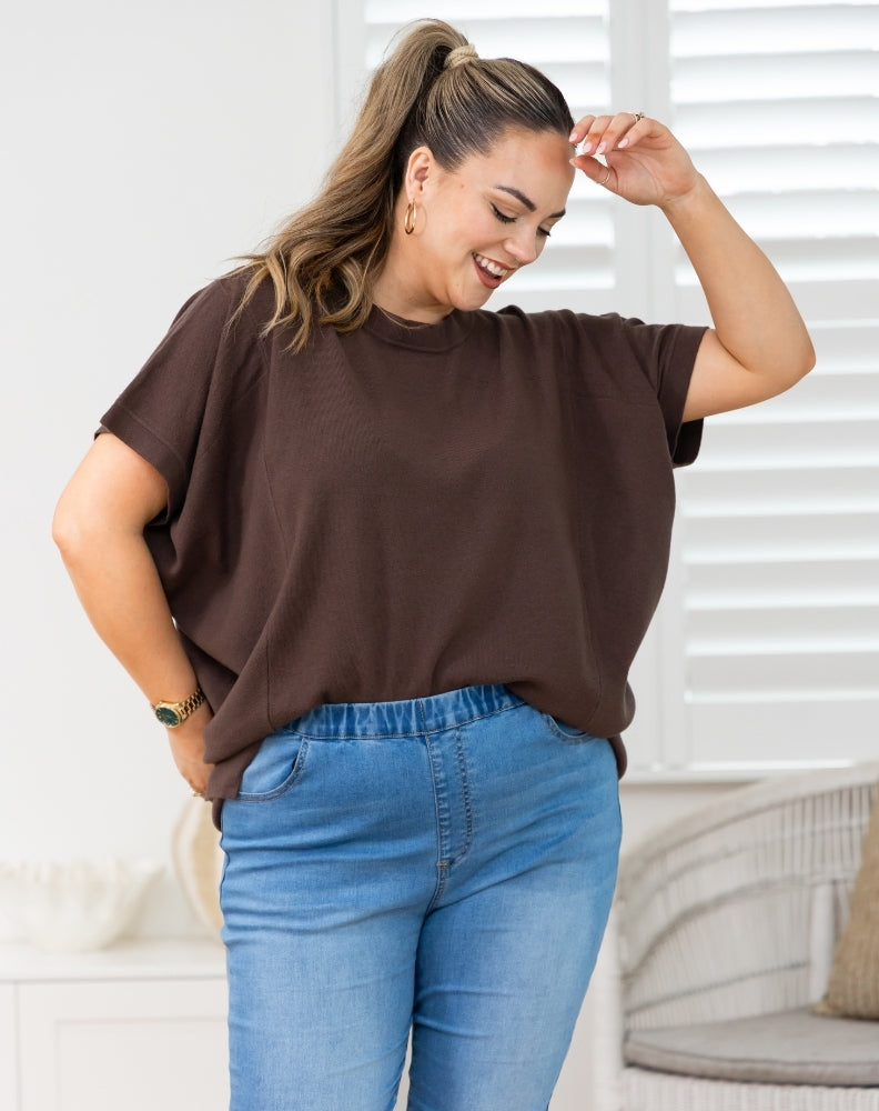 A woman wearing a loose-fitting brown short-sleeve t-shirt and blue jeans, posing in a brightly lit room with white window blinds in the background.