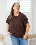A woman wearing an oversized brown short-sleeve t-shirt and blue jeans, standing in a brightly lit room with white shutters in the background.