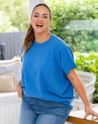 A woman wearing a bright blue short-sleeve crewneck t-shirt and blue jeans, smiling and posing in an outdoor patio setting with green foliage in the background.