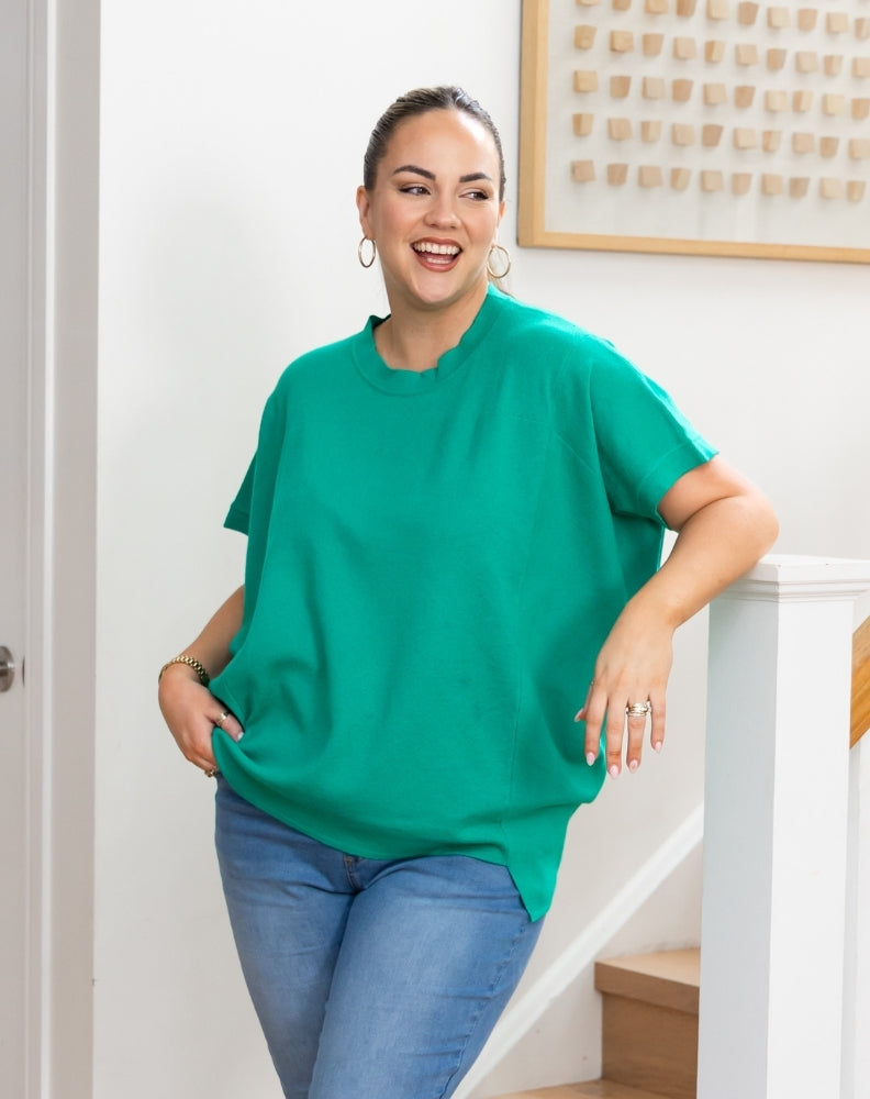 A woman wearing a bright green short-sleeve t-shirt and blue jeans, smiling and posing indoors next to a white staircase railing.