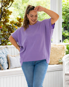 A woman wearing an oversized lavender-purple short-sleeve t-shirt and blue jeans, standing on a white porch with green foliage in the background.