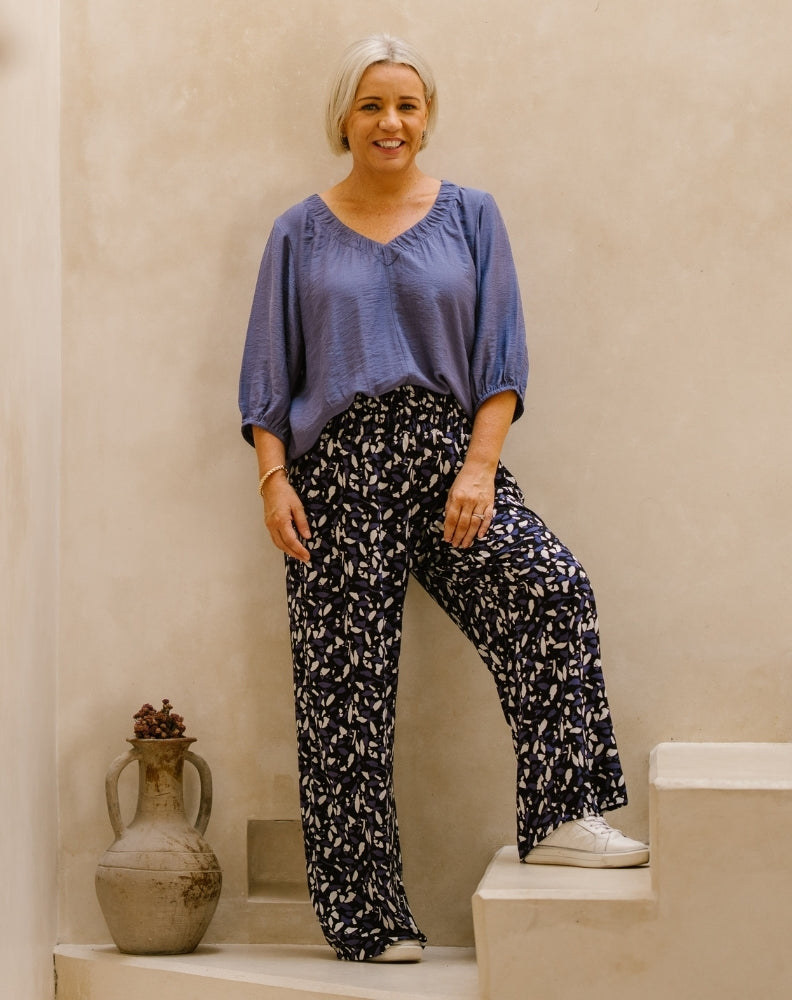 A woman with short blonde hair stands on stone steps wearing a loose blue blouse and dark navy wide-leg pants with a white abstract print. She is posed next to a rustic clay vase against a textured beige wall.