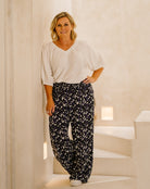 A woman stands indoors wearing a loose white blouse and dark patterned wide-leg pants with white shoes, posed against a warm beige wall and steps.