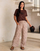 Woman wearing a brown t-shirt and patterned pants standing in a room with a neutral color scheme.