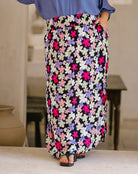 A person wearing a long black maxi skirt with a colorful floral print in pink, purple, white, and light blue, paired with a blue top and black sandals.